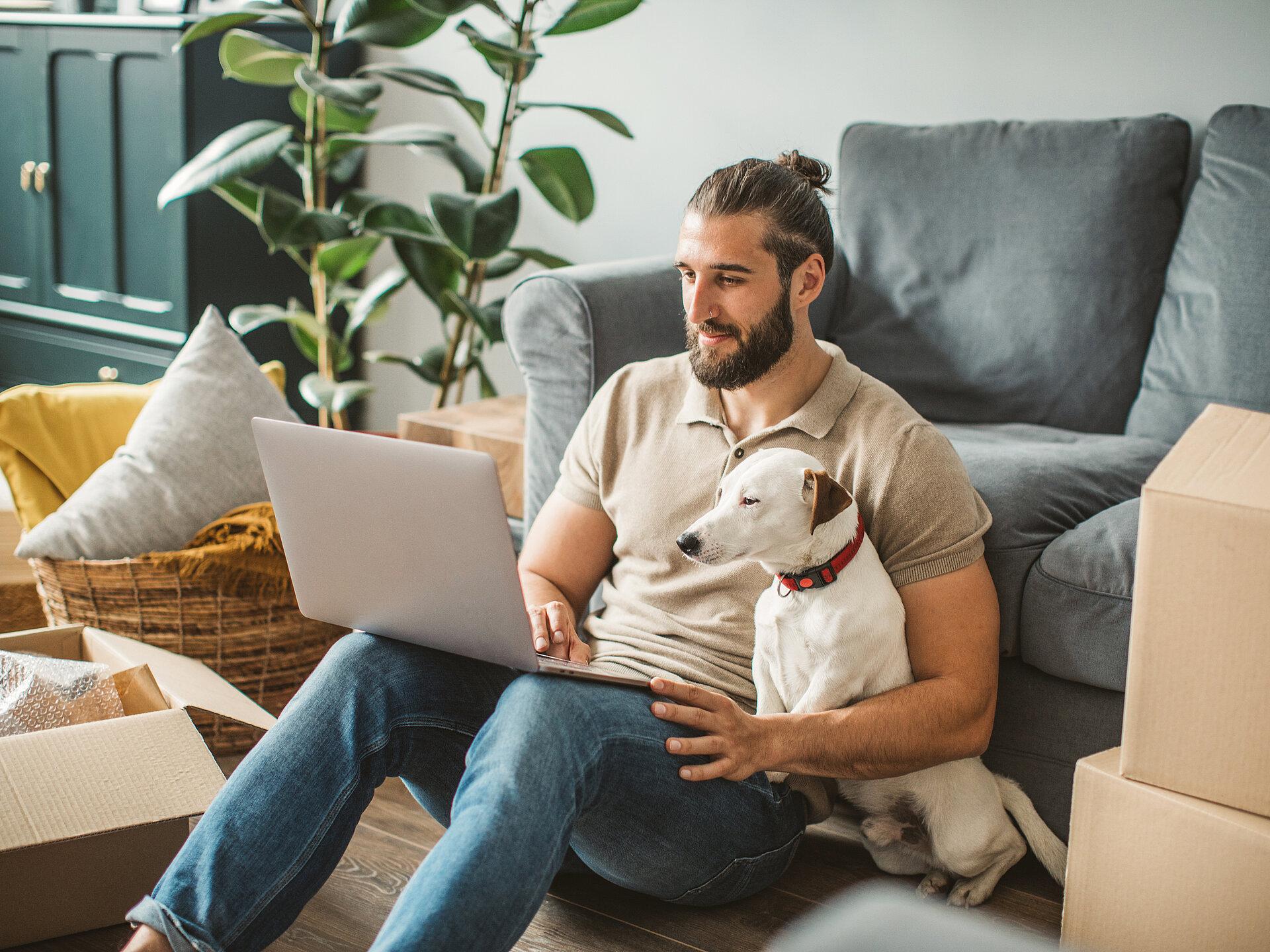 Ein Mann sitzt mit einem Hund auf dem Fußboden an eine Couch gelehnt und hat einen Laptop auf seinen Beinen