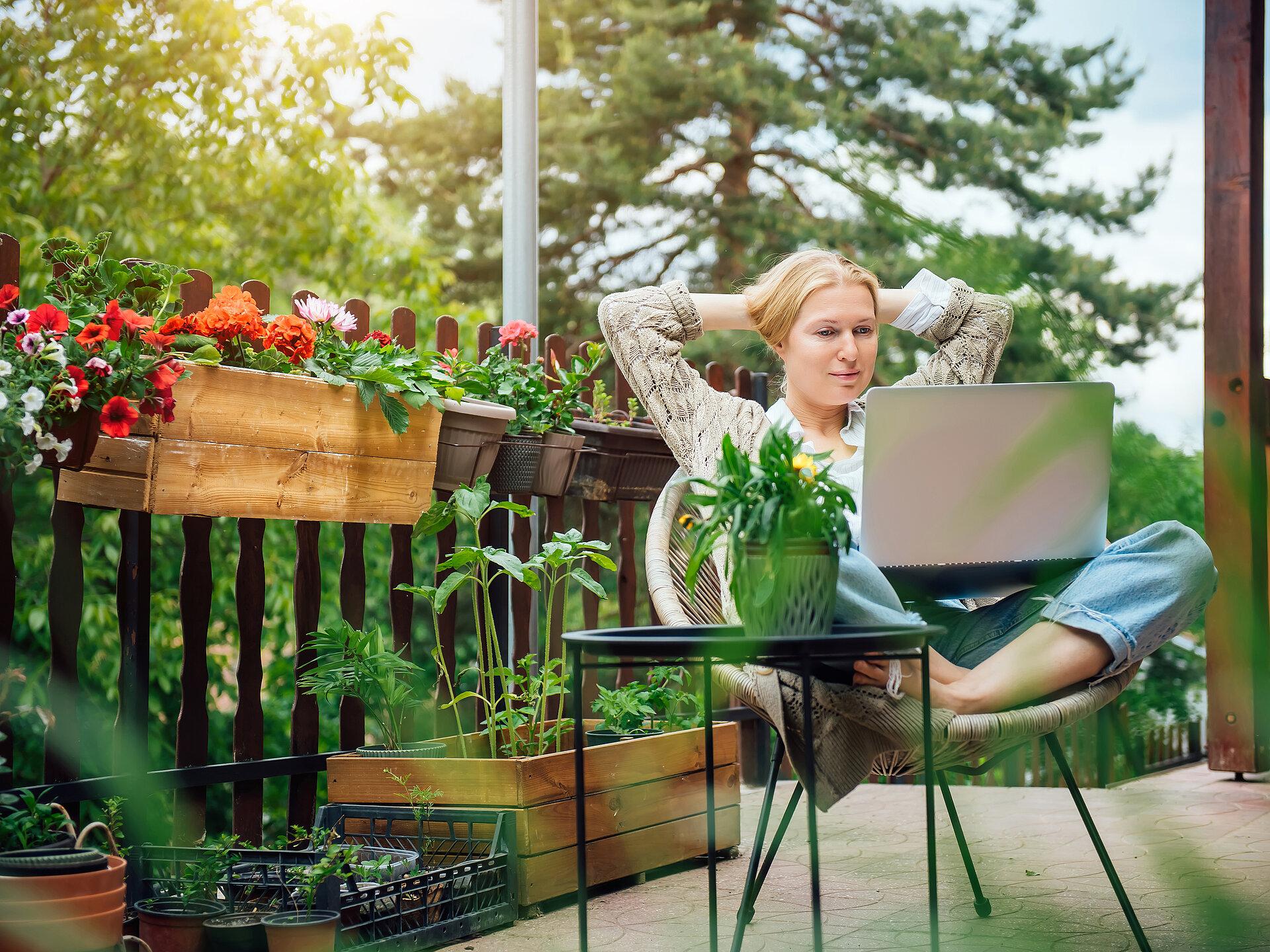 Eine blonde Frau mittleren Alters in ihrem Landhaus sitzt auf der Terrasse, umgeben von Blumen, und arbeitet an einem Laptop.