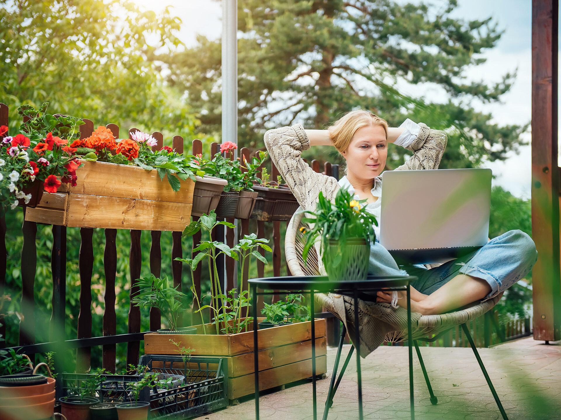 Eine blonde Frau mittleren Alters in ihrem Landhaus sitzt auf der Terrasse, umgeben von Blumen, und arbeitet an einem Laptop.