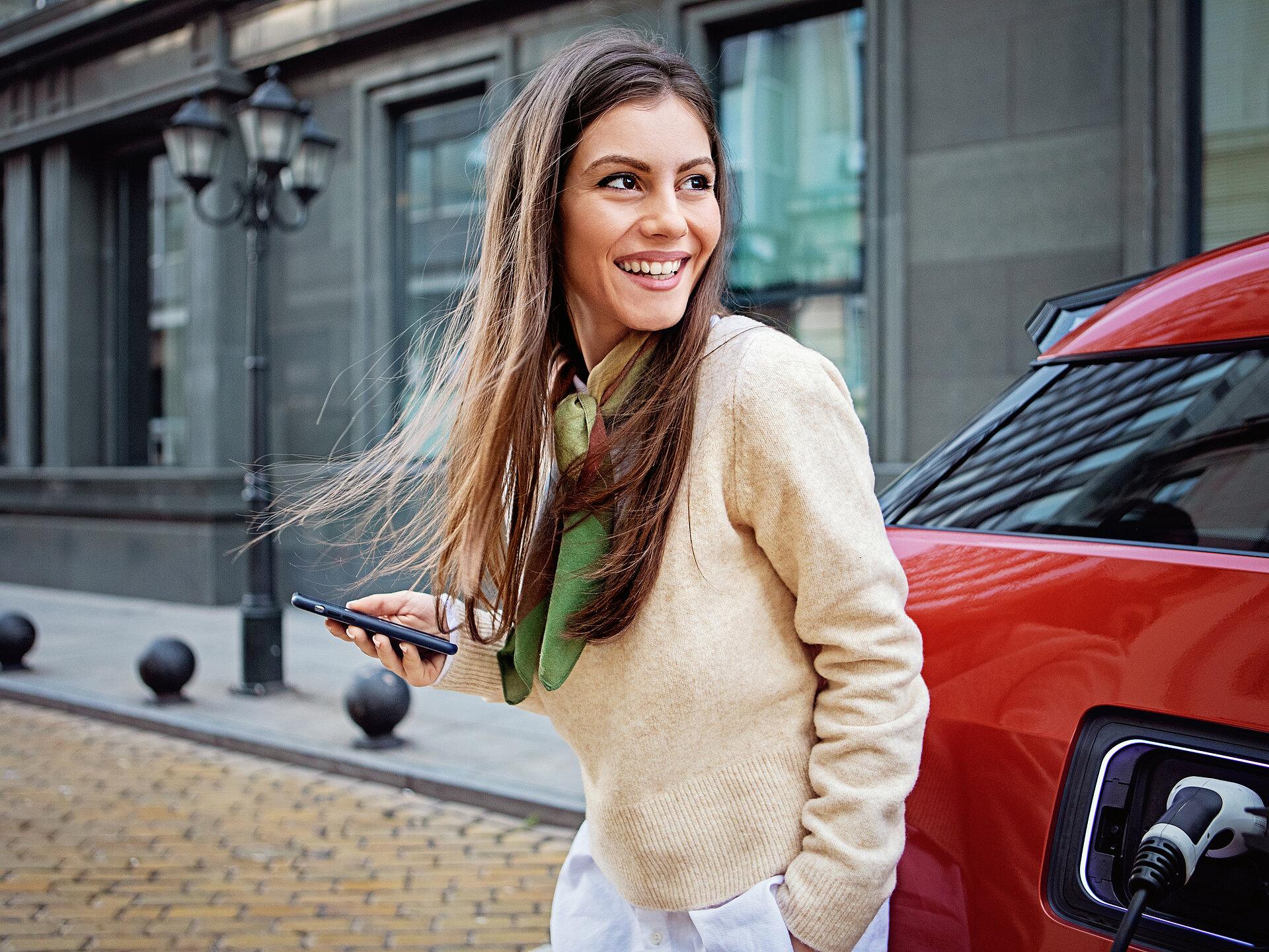 Eine lächelnde Frau steht vor einem roten E-Auto und hält ein Handy in der Hand. 