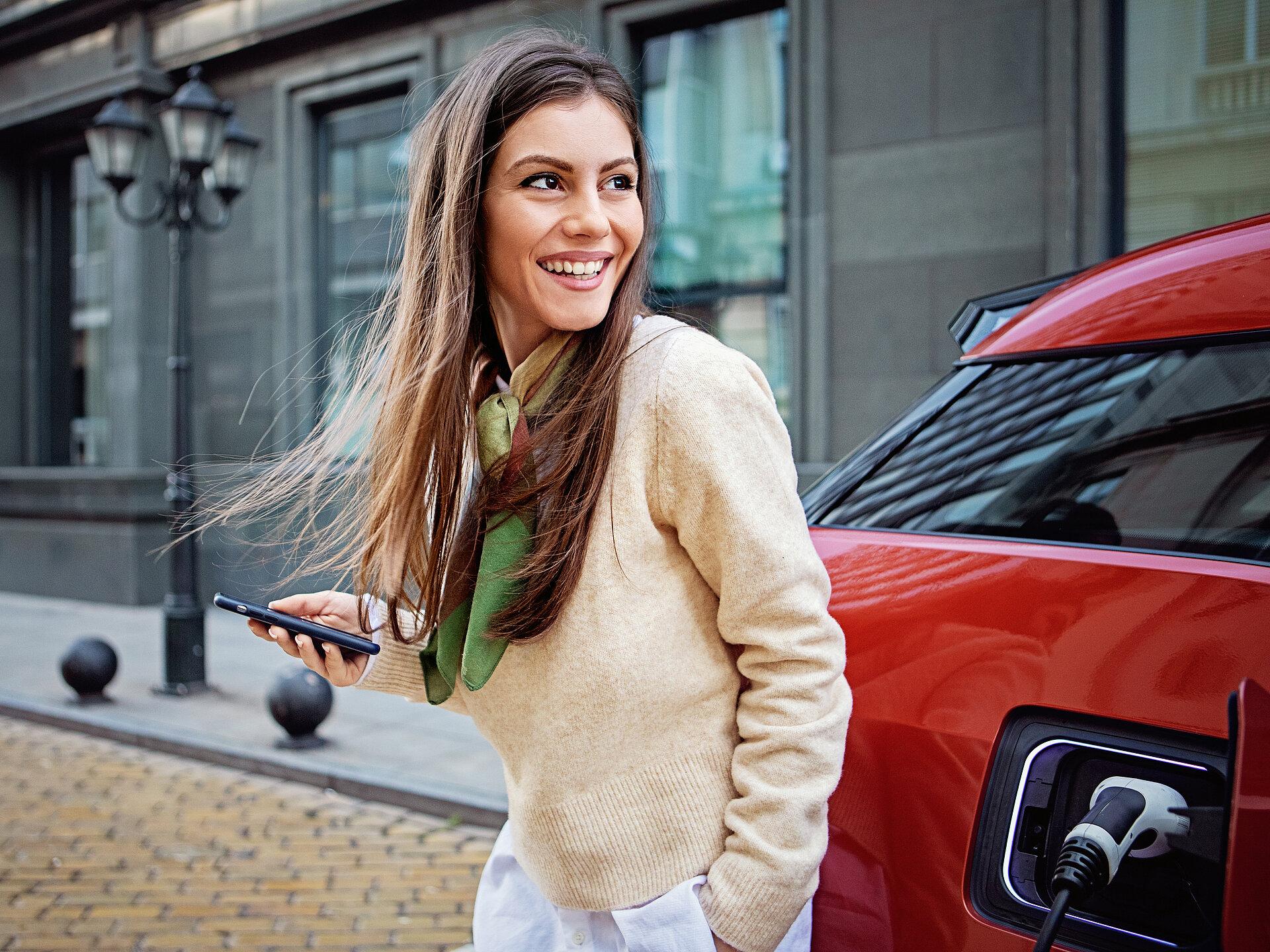 Eine lächelnde Frau steht vor einem roten E-Auto und hält ein Handy in der Hand. 