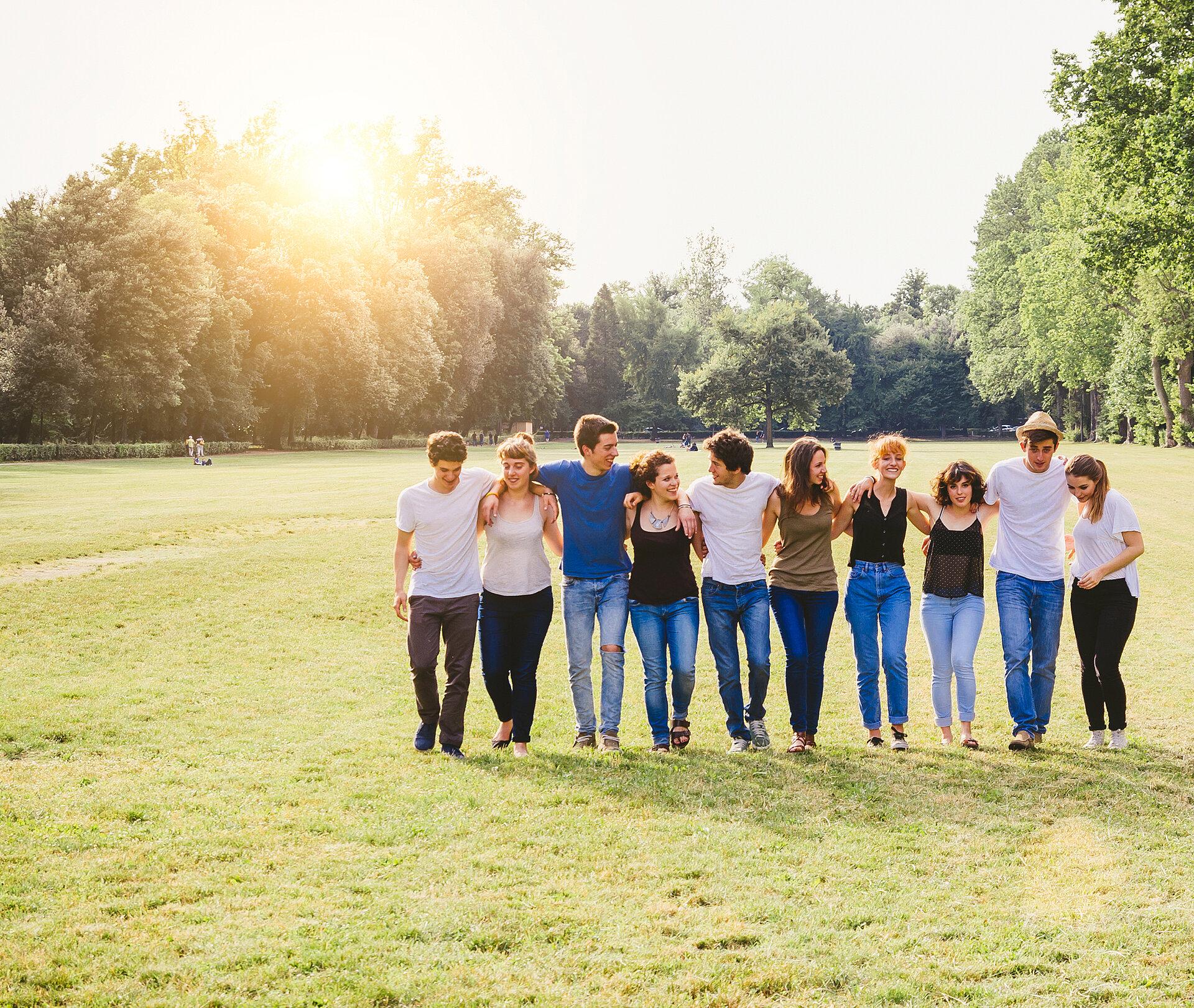 Zehn jungen Menschen stehen auf einer sonnigen Wiese in einer Parklandschaft in einer Reihe und ist gegenseitig eingehakt