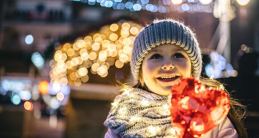 Lachendes kleines Mädchen mit Mütze draußen und einem roten Herzlolli in der Hand